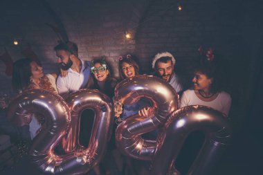 Bir grup genç arkadaş yeni yıl partisinde eğleniyor, ellerinde 2020 numara şeklinde balonlar var ve gece yarısı geri sayımında şampanya içiyorlar.