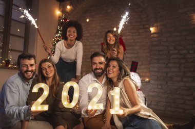 Yeni yıl partisinde eğlenen bir grup genç yeni yılı temsil eden 2021 numaralı aydınlatma numaralarını taşıyorlar ve gece yarısı havai fişeklerle el sallıyorlar.