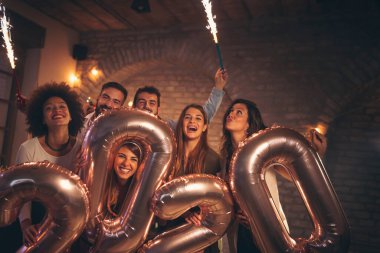 Bir grup genç arkadaş yeni yıl partisinde eğleniyor, 2020 numara şeklinde balonlar tutuyorlar ve gece yarısı geri sayımda maytaplarla el sallıyorlar.