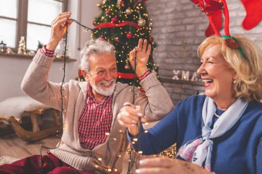 Güzel dekore edilmiş Noel ağacının yanında oturan neşeli yaşlı çift Noel için evi dekore ederken eğleniyor.
