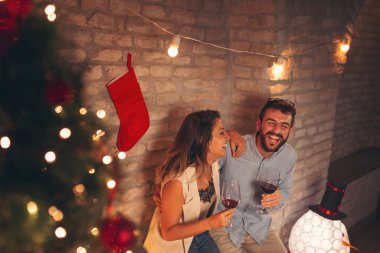 Güzel dekore edilmiş Noel ağacının yanında duran, şarap içen ve Noel 'i evde kutlarken eğlenen güzel genç çiftin yüksek açılı görüntüsü.