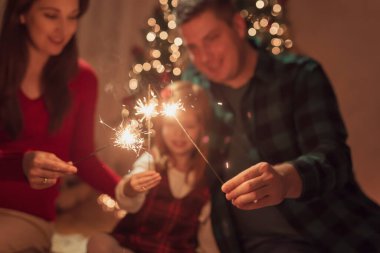Anne, baba ve kız Noel arifesini evde kutluyorlar, güzel dekore edilmiş Noel ağacının yanında yerde oturuyorlar, havai fişeklerle oynarken ve el sallarken eğleniyorlar.