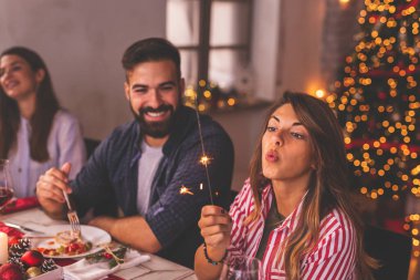 Bir grup neşeli genç arkadaş Noel yemeğinde eğleniyor, maytaplarla el sallıyor ve Noel şarkıları söylüyorlar.