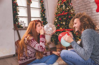 Birbirine aşık genç bir çift, Noel ağacının yanında oturmuş, Noel günü evde eğleniyor, balon üflüyor.