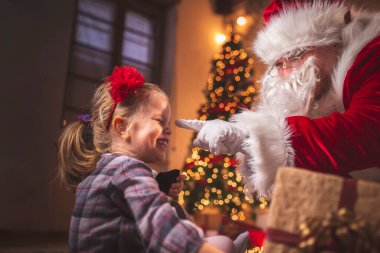 Güzel küçük kız Noel arifesinde evde eğleniyor, güzel dekore edilmiş bir Noel ağacının yanında oturuyor, Noel Baba gibi giyinmiş babasıyla Noel hediyelerini açıyor.