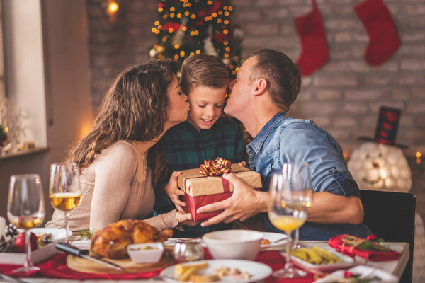 Семья с рождественским ужином дома, собрались вокруг стола, наслаждаясь их время вместе; мать и отец дарят рождественский подарок своему сыну и целует его