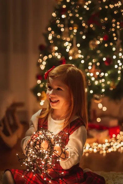 Güzel bir Noel ağacının yanında oturmuş Noel ışıklarını tutuyor ve düzenliyor, neşeyle oynuyor ve gülümsüyor.