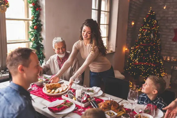 Mutlu bir aile, Noel yemeğini evde birlikte yiyor, birbiriyle konuşuyor ve gülüyor. Bu arada genç bir kadın da sofraya yemek koyuyor.