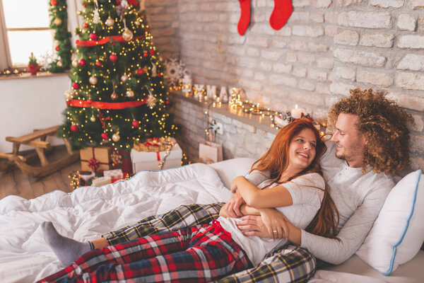 Beautiful young couple in love lying in bed on Christmas morning, hugging, cuddling and enjoying their time together during winter holiday season