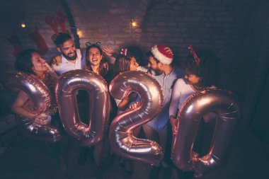 Bir grup genç arkadaş yeni yıl partisinde eğleniyor, ellerinde 2020 numara şeklinde balonlar var ve gece yarısı geri sayımında şampanya içiyorlar.