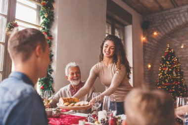Mutlu bir aile, Noel yemeğini evde birlikte yiyor, birbiriyle konuşuyor ve gülüyor. Bu arada genç bir kadın da sofraya yemek koyuyor.