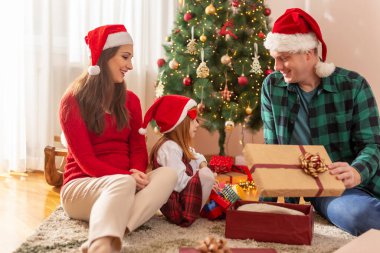 Güzel, genç bir aile Noel ağacının yanında oturuyor, Noel hediyelerini açarken eğleniyorlar, ebeveynler kızlarıyla oynuyorlar.