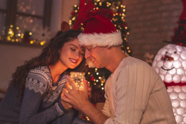 Genç aşık çift güzel dekore edilmiş Noel ağacının yanında oturuyor, kış tatilinin güzelliğinin tadını çıkarıyor, evde dinleniyor ve sarılıyor.