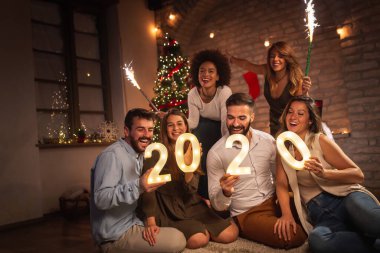 Yeni yıl partisinde eğlenen bir grup genç arkadaş, 2020 yılını temsil eden aydınlatıcı numaralar taşıyorlar ve gece yarısı havai fişeklerle el sallıyorlar.