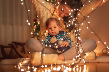 Anne oğlunu kucağında tutuyor, güzel süslenmiş Noel ağacının yanında oturuyor, yeni yılı kutluyor ve Noel ışıklarıyla oynuyor.