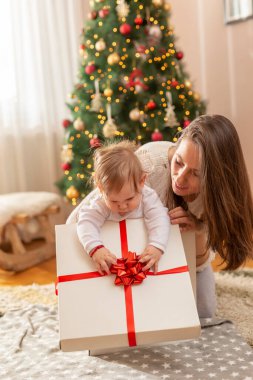 Anne ve bebek Noel günü evde eğleniyor, güzel dekore edilmiş Noel ağacının yanında oturuyor, hediyeleri açıyor ve oyuncaklarla, yaylarla ve kurdelelerle oynuyor.