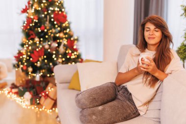Güzel genç bir kadın Noel günü evde dinleniyor, güzel dekore edilmiş Noel ağacının yanında kanepede oturuyor, kahve içiyor.