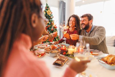 Bir grup neşeli genç arkadaş evde birlikte Noel yemeği yiyor, maytaplarla el sallıyor ve kadeh kaldırıyor.