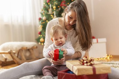 Anne ve oğlu Noel sabahı evde eğleniyor, güzel dekore edilmiş Noel ağacının yanında oturuyor, hediyeleri açıyor ve oyuncak kalıplarla oynuyor.