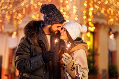 Birbirine aşık bir çift şehrin sokaklarında yeni yılı kutlarken geceyarısı geri sayımı için kıvılcımlar tutarken ve arka planda Noel ışıkları varken. Maytapa odaklan.
