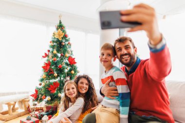 Güzel mutlu bir aile Noel 'i evde kutluyor, ebeveynler çocuklarla vakit geçiriyor, Noel ağacının yanında selfie çekiyor.
