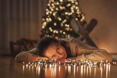 Güzel genç bir kadın güzel dekore edilmiş Noel ağacının yanında yerde uzanıyor, evde dinleniyor ve kış hologünü sezonunun tadını çıkarıyor.