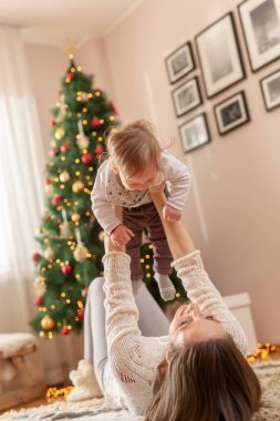 Anne Noel sabahı bebeğiyle oynarken eğleniyor, yere uzanmış, güzelce süslenmiş Noel ağacının yanında, onu kaldırıyor ve gülüyor.