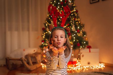 Geyik boynuzu takan güzel bir kız Noel ağacının yanında oturmuş Noel arifesini evde kutlarken konfeti üflerken eğleniyor.