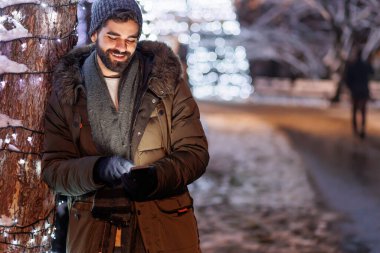 Noel arifesini şehir sokaklarında Noel ışıklarıyla geçirirken neşeli, yakışıklı genç adam akıllı telefon kullanıyor.