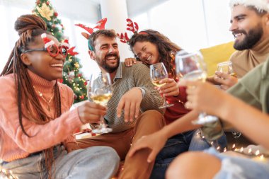 Bir grup neşeli genç arkadaş güzelce süslenmiş Noel ağacının yanında oturmuş Noel 'i evde kutluyor, şarap içiyorlar.