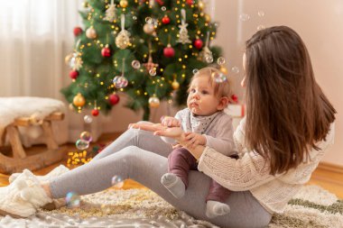 Genç anne Noel sabahı bebeğiyle oynuyor, onu kucağında tutuyor, sabun köpükleri yapıyor ve yakalıyor, güzelce süslenmiş Noel ağacının yanında oturuyor.