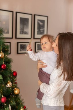 Anne oğlunu kollarında tutuyor ve ilk Noel ağacını süslerken eğleniyor.