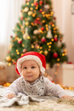 Noel Baba şapkası takmış güzel bir Noel ağacının yanında uzanmış, Noel sabahı gülümseyen güzel bir çocuk.