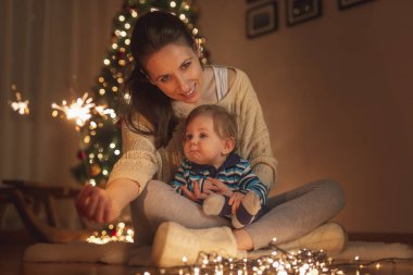 Anne oğlunu kucağında tutuyor, Noel arifesini evde kutluyor, güzelce süslenmiş Noel ağacının yanında oturuyor, maytaplarla oynuyor.