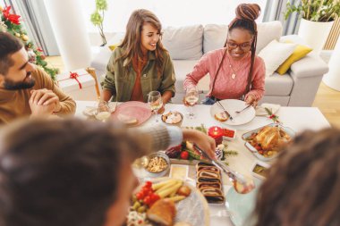 Bir grup neşeli genç arkadaş evde birlikte Noel yemeği yiyor, yemek tabaklarını birbirlerine dağıtıyor ve sohbet ediyorlar.