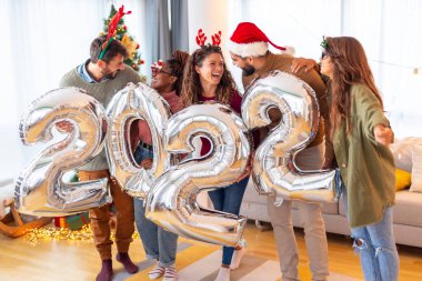 Bir grup genç arkadaş, 2022 numaralı dev balonları tutarak evde yeni yılı kutluyor.