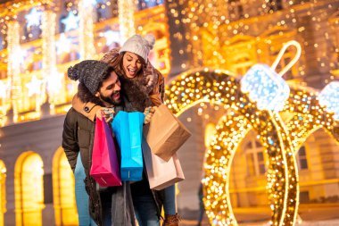 Genç ve güzel bir çift Noel alışverişi yaparken eğleniyor. Erkek, Noel ışıklarıyla alışveriş torbaları taşırken kadını sırtında taşıyor.