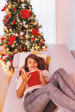 Noel arifesinde evde dinlenen, Noel ağacının yanındaki kanepede uzanmış kitap okuyan, kış tatilinin tadını çıkaran güzel genç bir kadın.