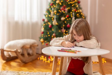 Güzel küçük kız kış tatilinin tadını çıkarıyor, güzel dekore edilmiş Noel ağacının yanında oturuyor, pastel boyalarla resim çizerken eğleniyor.