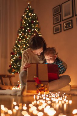 Güzel anne küçük oğlunu kucağında tutuyor, güzel dekore edilmiş Noel ağacının yanında oturuyor ve sihirli, parıldayan Noel hediyesini açarken eğleniyor.