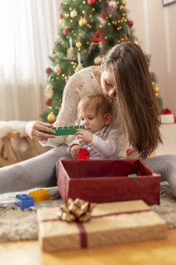 Anne ve oğlu Noel sabahı evde eğleniyor, güzel dekore edilmiş Noel ağacının yanında oturuyor, hediyeleri açıyor ve oyuncak kalıplarla oynuyor.