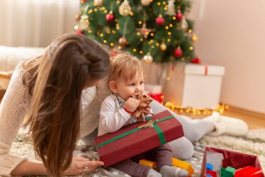 Anne ve bebek Noel günü evde eğleniyor, güzel dekore edilmiş Noel ağacının yanında oturuyor, hediyeleri açıyor ve oyuncaklarla, yaylarla ve kurdelelerle oynuyor.