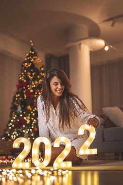 Bir kadın Noel ağacının yanında yerde oturuyor, 2022 yılını aydınlatıyor ve evde yeni yılı kutluyor.