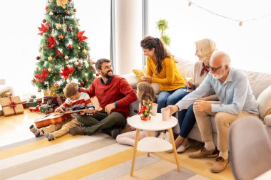 Çok nesildir Noel 'i evde birlikte geçirip eğlenen aile Noel ağacının etrafında toplanmış gitar çalıyor ve şarkı söylüyor. 