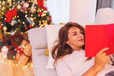 Noel arifesinde evde dinlenen, Noel ağacının yanındaki kanepede uzanmış kitap okuyan, kış tatilinin tadını çıkaran güzel genç bir kadın.