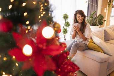 Güzel genç bir kadın Noel günü evde dinleniyor, Noel ağacının yanındaki kanepede oturuyor, kahve içiyor.