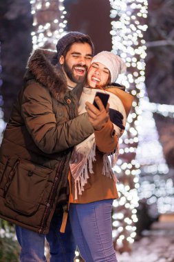 Güzel, genç ve aşık bir çift, arka planda bir sürü Noel ışıklarıyla şehrin sokaklarında yılbaşı arifesini kutlarken akıllı telefonlar kullanarak selfie çekerek eğleniyor.