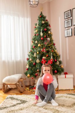 Geyik boynuzu takan küçük bir kız Noel ağacının yanında oturmuş Noel 'i evde kutlarken balon şişirip eğleniyor.