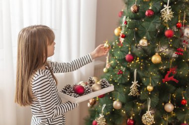 Güzel küçük kız Noel gününü evde, Noel ağacını ve kış tatili için evini süsleyerek geçiriyor.