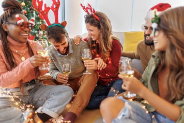 Bir grup neşeli genç arkadaş güzelce süslenmiş Noel ağacının yanında oturmuş Noel 'i evde kutluyor, şarap içiyorlar.